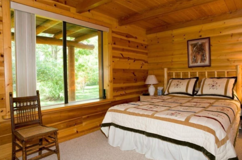 Cozy wooden bedroom with a bed, decorative quilt, chair, and large window overlooking a green outdoor view.