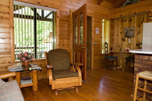 Cozy wooden interior of a cabin with a rocking chair, plants, and a dining area visible in the background.