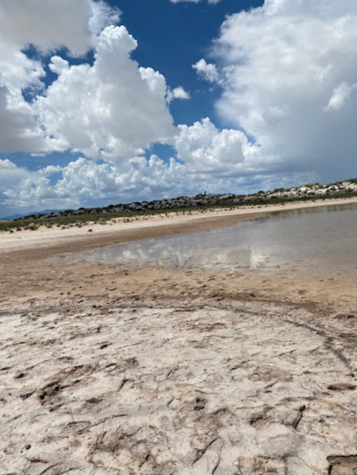 A dry, cracked landscape with a shallow pool of water reflecting fluffy clouds and blue sky.