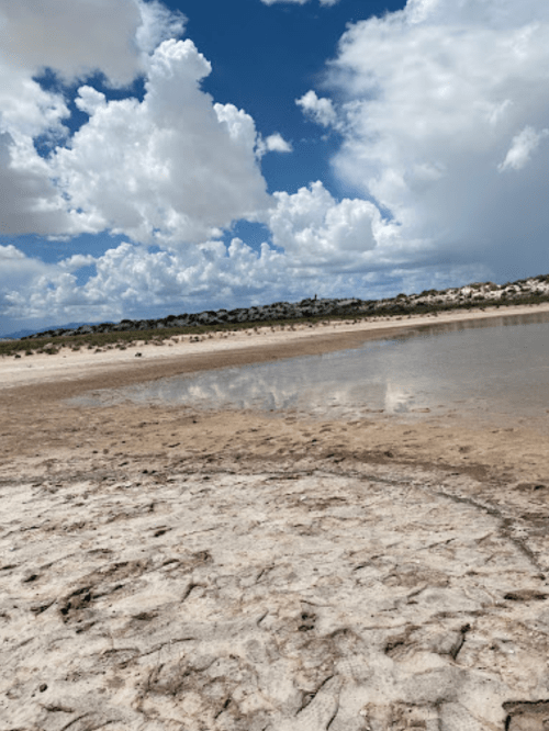 A dry, cracked landscape with a shallow pool of water reflecting fluffy clouds and blue sky.