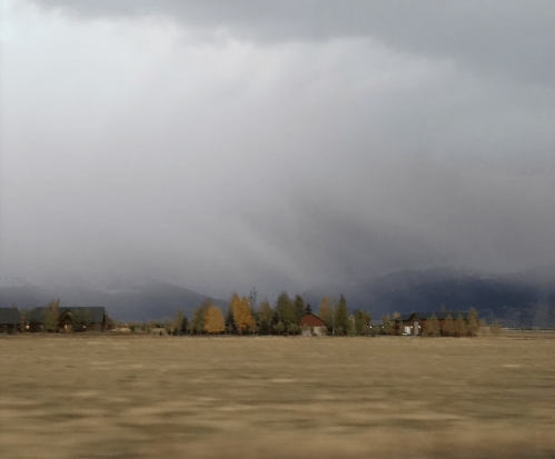 A cloudy sky looms over a rural landscape with distant mountains and scattered trees, hinting at an approaching storm.