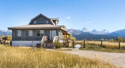 A rustic house surrounded by tall grass, with mountains in the background under a clear blue sky.