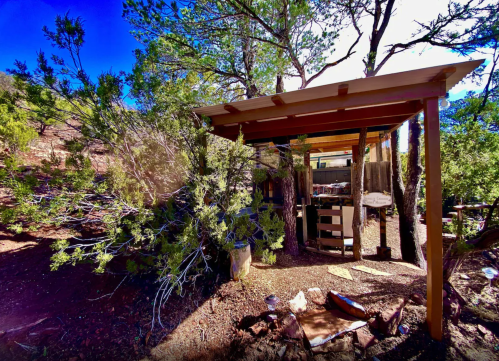 A rustic outdoor structure surrounded by trees, with a glimpse of furniture and a natural landscape in the background.