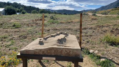 A topographical model of a landscape, set on a wooden stand, with hills and vegetation in the background.