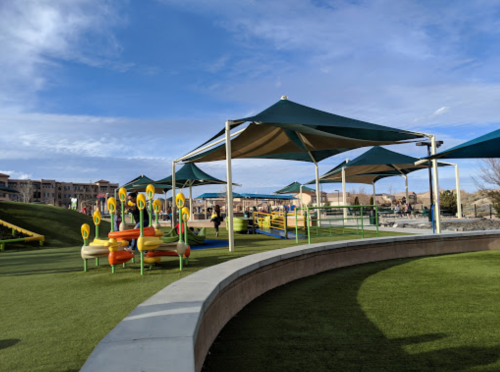 A colorful playground with shaded areas, green grass, and modern equipment under a blue sky.