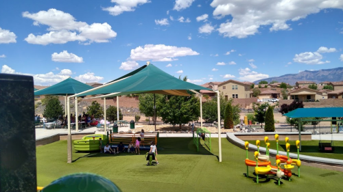 A playground with green turf, shaded areas, and colorful equipment under a blue sky with fluffy clouds.