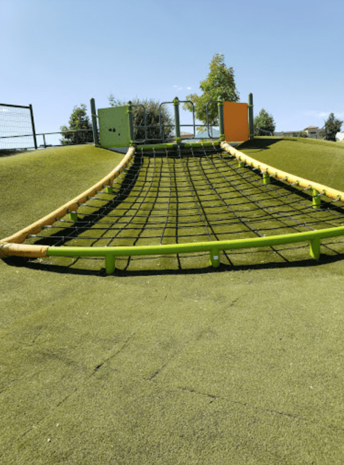 A playground climbing structure with a net on a grassy hill under a clear blue sky.