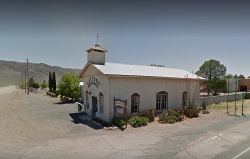 A small, white chapel with a cross on top, surrounded by trees and gravel, set against a mountainous backdrop.
