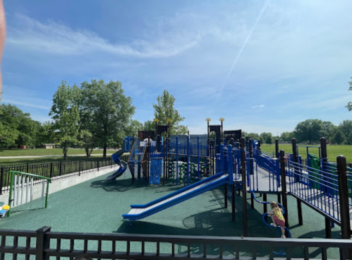 A colorful playground with slides and climbing structures, surrounded by green grass and trees under a clear blue sky.