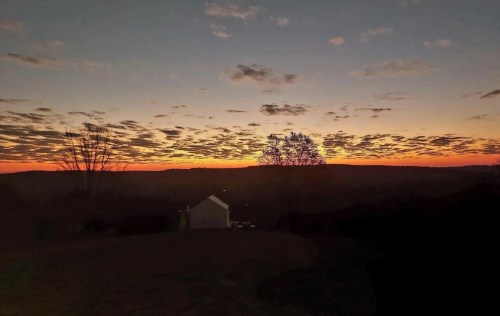 A serene sunset over rolling hills, with a silhouette of a tree and a small building in the foreground.