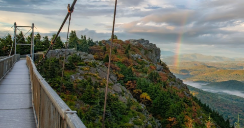 A scenic view from a suspension bridge overlooking mountains and a rainbow in the distance, surrounded by autumn foliage.