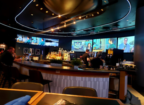 A modern bar with multiple screens showing people, featuring a well-stocked counter and patrons seated at tables.