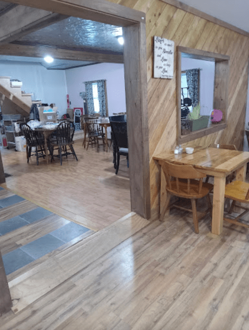 A cozy dining area with wooden tables and chairs, featuring a warm, rustic design and a view into another room.