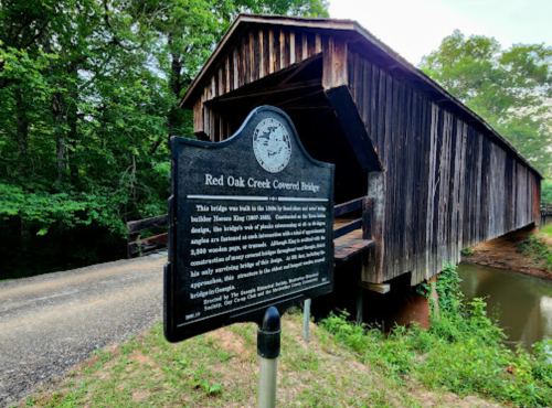 A historic sign in front of the Red Oak Creek Covered Bridge, surrounded by trees and a calm waterway.
