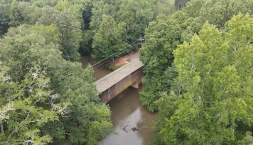 Aerial view of a wooden covered bridge over a river, surrounded by lush green trees.