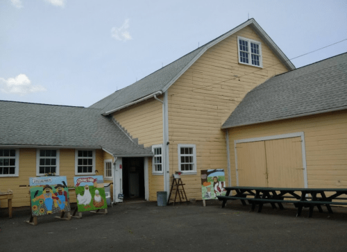 A yellow barn with a gray roof, featuring colorful murals and picnic tables outside.