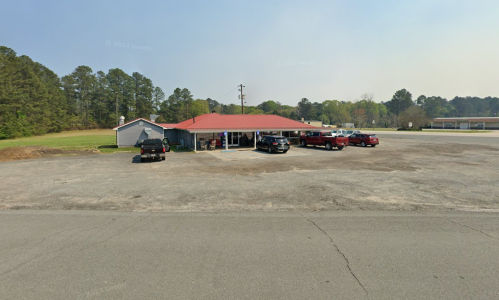 A one-story building with a red roof, surrounded by parked cars, set in a rural area with trees in the background.
