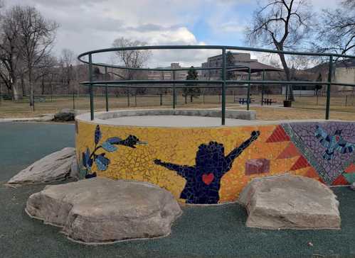 Colorful mosaic wall with a silhouette of a child, surrounded by rocks in a park setting with trees and buildings in the background.