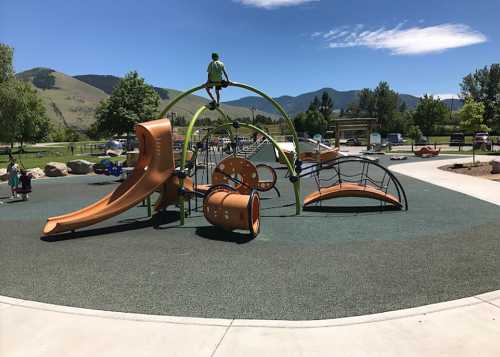 A colorful playground with slides and climbing structures, set against a backdrop of green hills and blue sky.