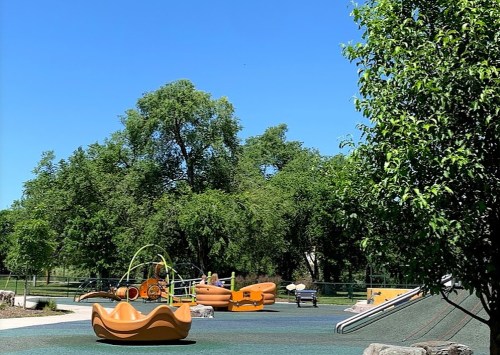 A sunny playground with colorful equipment, including slides and climbing structures, surrounded by green trees.