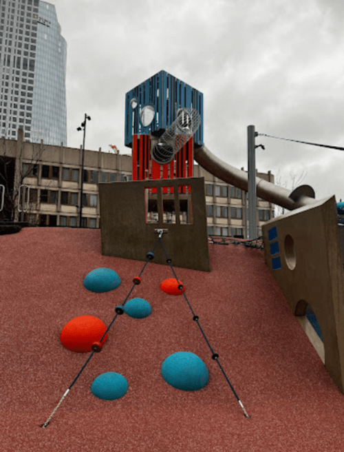 A colorful playground with a climbing structure, red and blue shapes, and a slide, set against a cloudy sky.