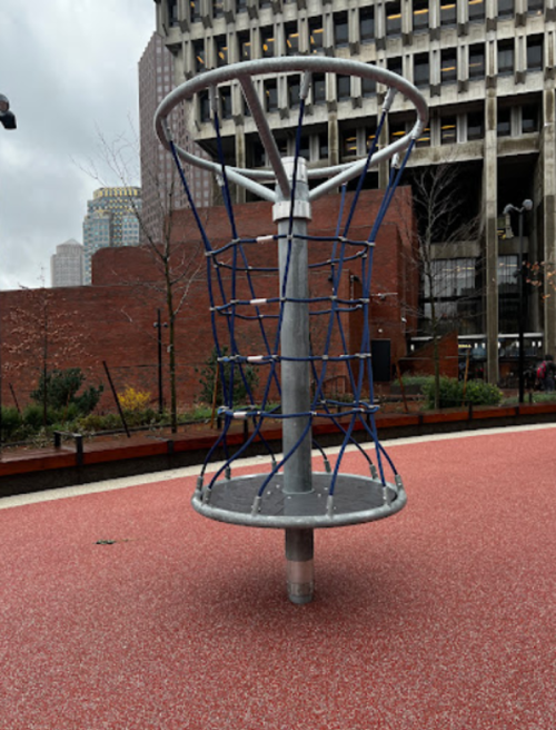 A metal climbing structure with blue ropes, set on a red rubber surface in an urban playground.