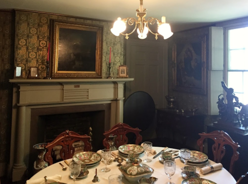 A vintage dining room with a set table, ornate chairs, a fireplace, and framed artwork on the walls.