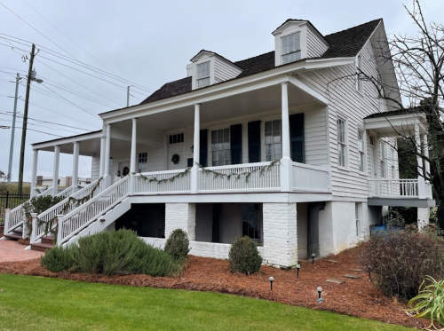 A large white house with a porch, surrounded by greenery and a landscaped yard, under a cloudy sky.