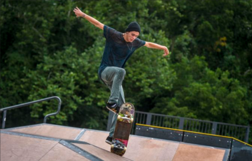 A skateboarder performs a trick on a ramp, surrounded by greenery in the background.