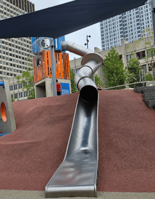 A large metal slide descends from a colorful playground structure, surrounded by red rubber mulch and city buildings.