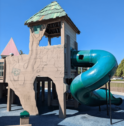 A playground structure with a green slide and a wooden playhouse design under a clear blue sky.