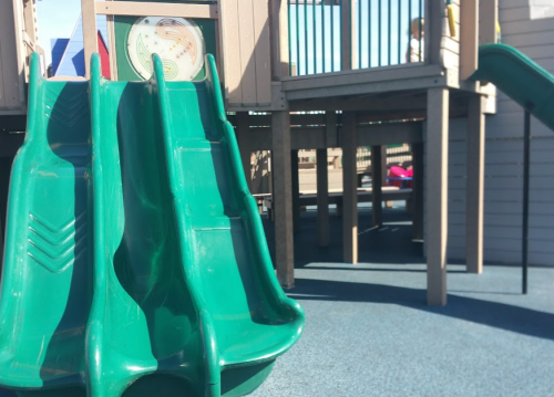 A green slide at a playground, with a wooden structure and blue rubber flooring in the background.
