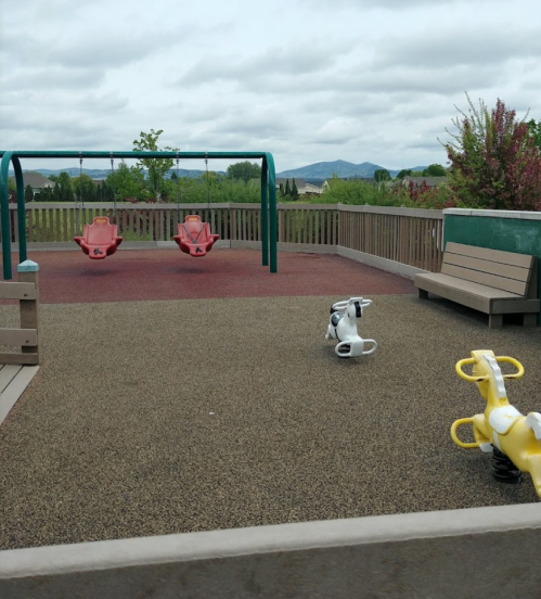Empty playground with swings, benches, and spring riders on a rubber surface under a cloudy sky.