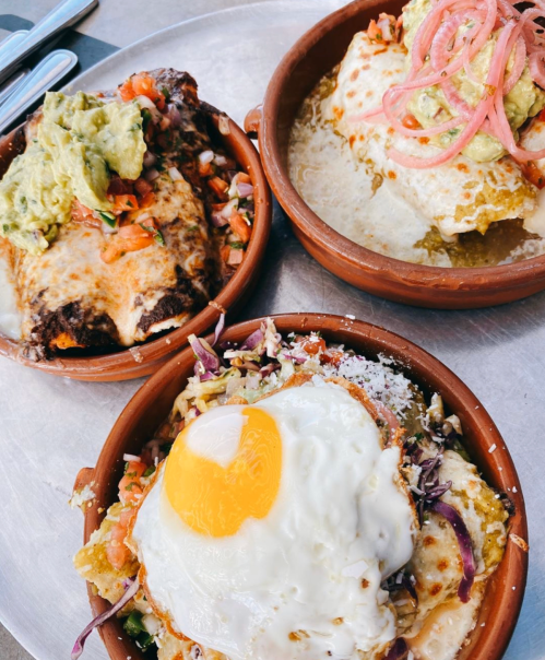 Three clay dishes with colorful Mexican dishes: enchiladas, guacamole, pico de gallo, and a fried egg on top.