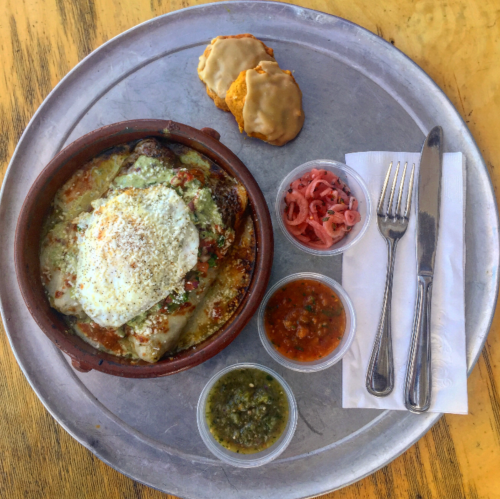 A metal tray with a clay dish of layered food topped with cheese, alongside salsas, pickled onions, and cookies.
