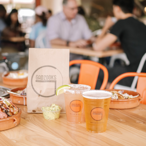A table with two drinks, a paper bag labeled "Gadzooks," and food in bowls, set in a busy dining area.