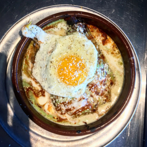 A baked dish with a sunny-side-up egg on top, surrounded by creamy sauce and green ingredients in a brown bowl.