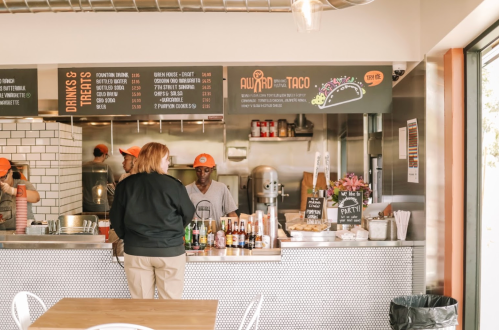 A bright taco restaurant interior with a menu board, staff serving, and a customer at the counter.