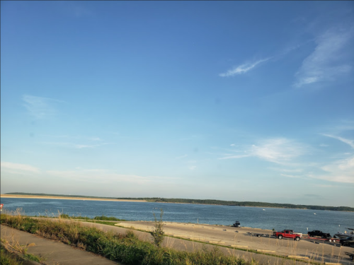 A serene lakeside view with clear blue skies and a distant shoreline, featuring parked boats and vehicles.