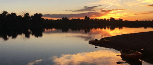 A serene sunset over a calm river, with silhouettes of trees and kayaks on the shore reflecting in the water.