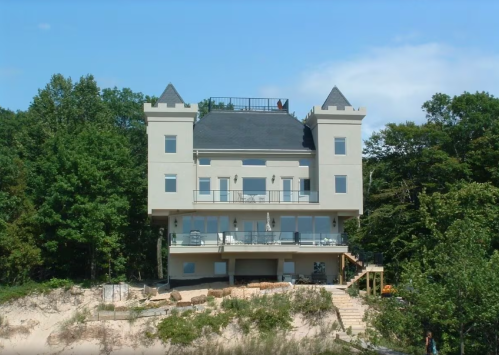 A large, modern house resembling a castle, surrounded by trees and situated on a sandy area.