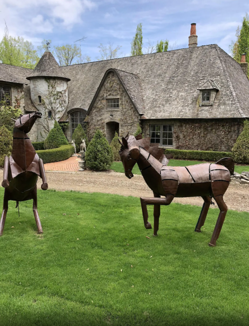 Two metal horse sculptures stand on green grass in front of a stone house surrounded by trees and shrubs.