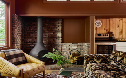 Cozy living room with a brick fireplace, yellow armchair, patterned sofa, and a fern plant. Warm, inviting atmosphere.