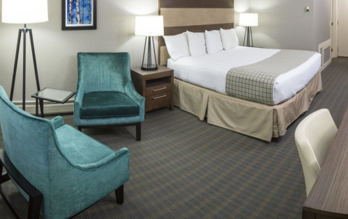 A modern hotel room featuring a king-sized bed, two lamps, a teal armchair, and a wooden desk.