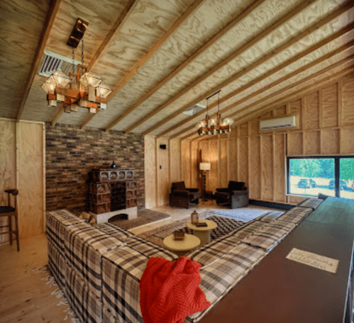 Cozy living room with wooden walls, a stone fireplace, and a plaid sofa, featuring warm lighting and a rustic decor.