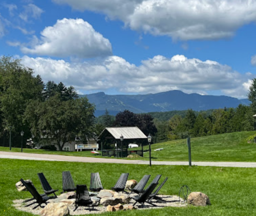 A scenic view of a fire pit surrounded by chairs, with mountains and a blue sky in the background.