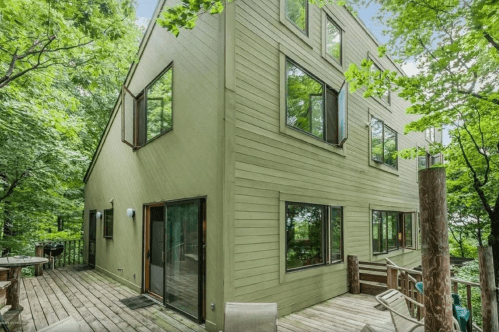 A modern green house surrounded by trees, featuring large windows and a wooden deck.