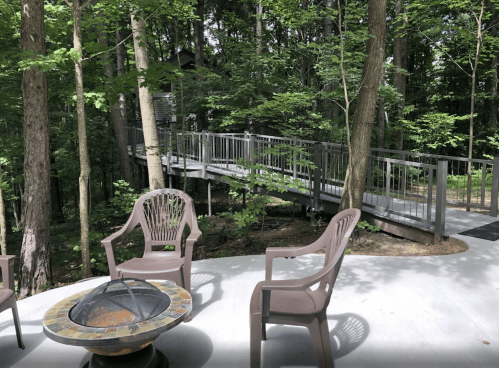 A serene outdoor space with four chairs around a fire pit, surrounded by trees and a wooden walkway.