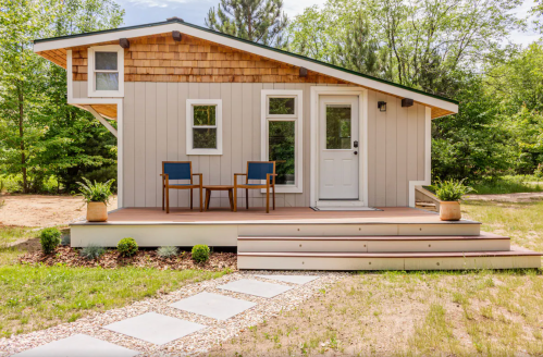 A modern small house with a wooden deck, two chairs, and a landscaped yard surrounded by trees.