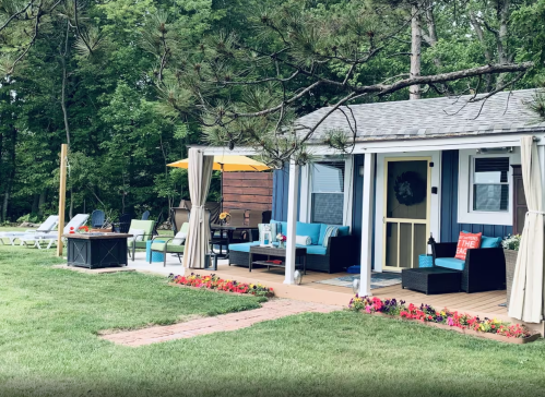 A cozy cottage with a porch, surrounded by greenery and colorful flowers, featuring outdoor seating and a fire pit.